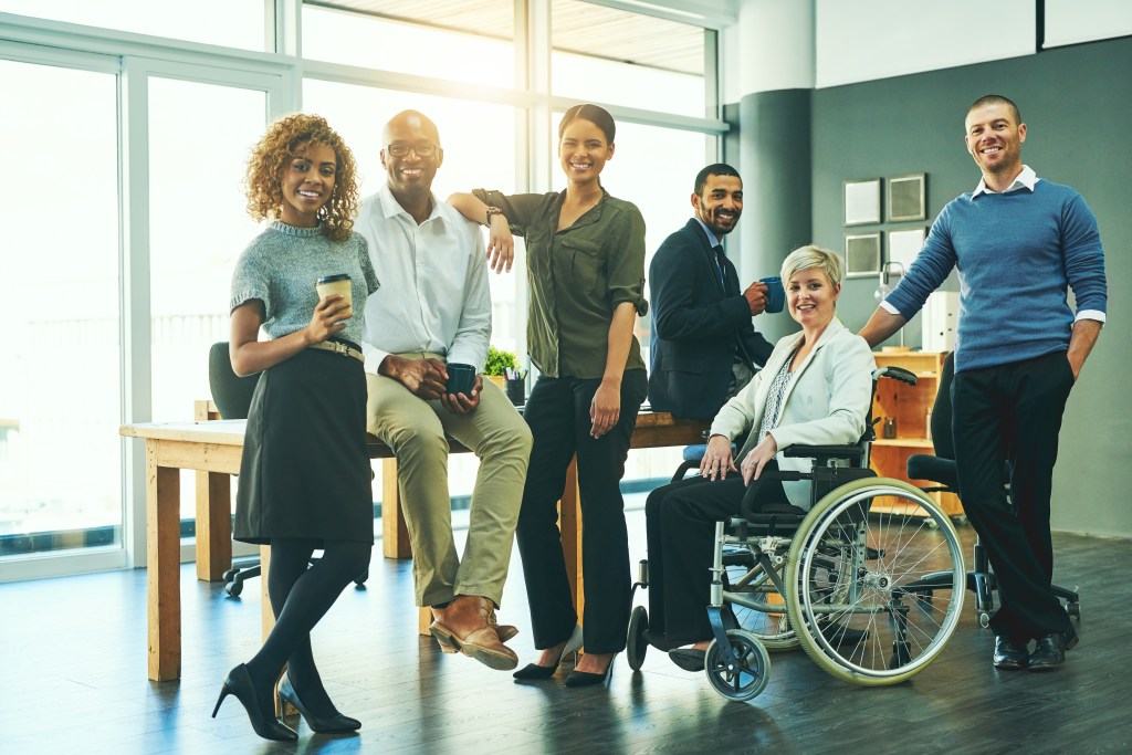 An ethnically diverse group of happy individuals with three individuals standing, two sitting on a table and one sitting in a wheelchair.